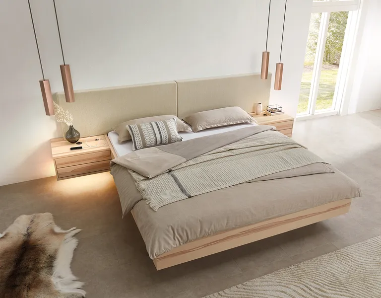 Schlafzimmer mit Massivholzbett, beige gepolstertem Kopfteil und integrierten Nachttischen, kombiniert mit warmen Pendelleuchten und natürlicher Bettwäsche für ein beruhigendes Schlafambiente.